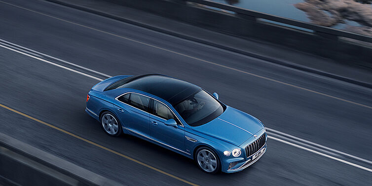 Thomas Exclusive Cars GmbH New Bentley Flying Spur Azure sedan in Light windsor blue paint by Mulliner, with 22 inch Azure wheels seen from overhead driving dynamically on a country road