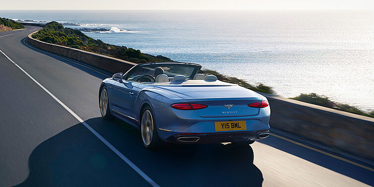 Thomas Exclusive Cars GmbH New Bentley Continental GTC Azure convertible rear view in Light Windsor blue paint with 22 inch ten spoke wheels driving dynamically on an ocean road in the sunshine